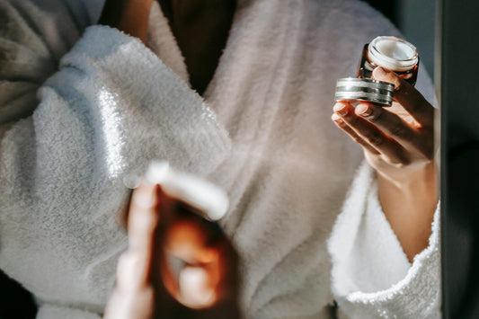 Woman in front of mirror wearing a bathrobe, applying eye cream.
